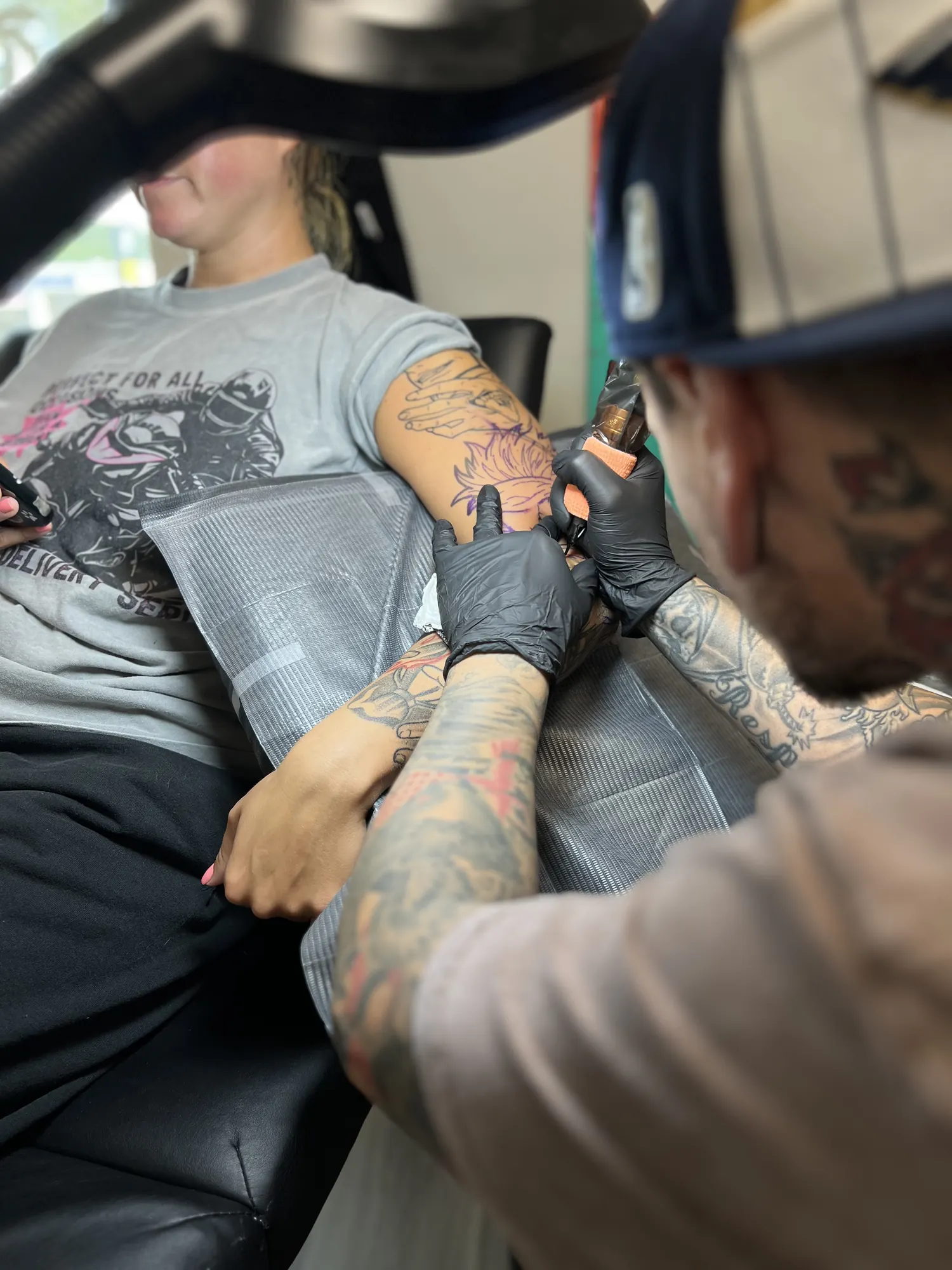 Cory Veron in a cap and black gloves, tattooing a client's upper arm